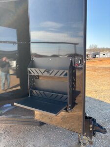 A black metal fold-down work table is mounted on the front of a glossy black Alumi Tech horse trailer parked on gravel. The table is unfolded, with other trailers and orange dirt visible in the background. -Stillwater Trailer Sales