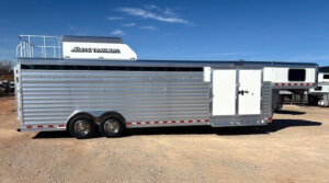 A large silver gooseneck livestock trailer with ribbed sides and a raised front section, featuring Horse Smart Tack storage, is parked on gravel under a clear blue sky. -Stillwater Trailer Sales