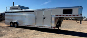 A large, silver Elite 6 livestock trailer with multiple windows and double axles is parked on a gravel lot under a clear blue sky, perfect for any Horse Smart Tack needs. Tack #23223. -Stillwater Trailer Sales