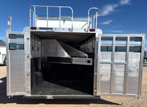 Open silver livestock trailer with rear doors open, showing interior divided by angled metal gates and partitions for transporting animals. Parked on a dirt lot under a clear blue sky, this trailer is perfect for Horse Smart Tack or Elite Horse Tack needs. -Stillwater Trailer Sales