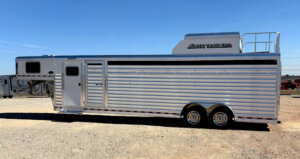 A large silver Elite 6 Horse livestock trailer with a gooseneck hitch is parked on gravel under a clear blue sky. It features dual axles, ventilation slats, and a roof rack labeled Elite Trailers, perfect for Horse Smart Tack needs. -Stillwater Trailer Sales