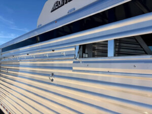Close-up of a shiny silver Horse Smart Tack Elite 6 Horse trailer with an open side vent, reflecting sunlight. The trailer has horizontal ridges and a partial logo visible at the top, set against a blue sky background. -Stillwater Trailer Sales