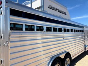 A shiny silver Elite 6 Horse livestock trailer with horizontal ventilation slats, double axles, and Horse Smart Tack storage, plus the brand name Elite Trailers visible on the upper section under a clear blue sky. -Stillwater Trailer Sales