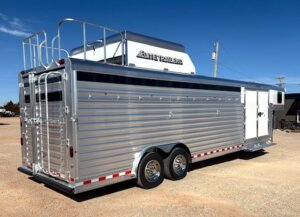 An Elite 6 large silver livestock trailer with ribbed metal sides, dual axles, rooftop ventilation, and a rear swing door is parked on a gravel lot under a clear blue sky&mdash;perfect for hauling Horse Tack or Smart Tack. -Stillwater Trailer Sales