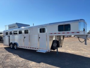 A large, silver Elite 5H gooseneck horse trailer with side load access, multiple windows and vents is parked on gravel under a clear blue sky. The 413 trailer features dual axles, a ramp, and convenient side doors. -Stillwater Trailer Sales