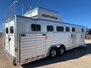 A large silver Elite 5H horse trailer with four windows, side vents, and a roof storage area is parked on dirt under a clear blue sky. This spacious Side Load trailer is marked with the brand Elite Trailers. -Stillwater Trailer Sales