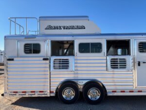A silver Elite 5H Trailers horse trailer with multiple windows and vents, side load access, two wheels visible, and a rooftop area against a clear blue sky. The trailer is parked on a gravel surface. -Stillwater Trailer Sales