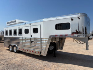 A large silver and white Bloomer 4H Trainer gooseneck horse trailer with multiple windows is parked on a gravel lot under a clear blue sky. -Stillwater Trailer Sales