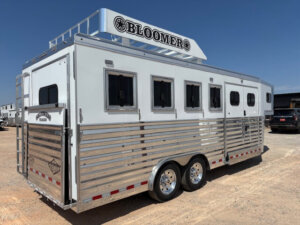 A large white and silver Bloomer 4H Trainer horse trailer with four windows and dual axles is parked on dirt under a clear blue sky. The trailer features a roof rack and shiny metal siding. -Stillwater Trailer Sales