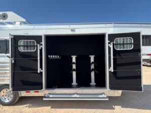 Open Bloomer horse trailer tack room with both doors wide, showing interior saddle racks, bridle hooks, and carpeted walls. The metallic silver Trainer 549 exterior gleams under a clear blue sky. -Stillwater Trailer Sales