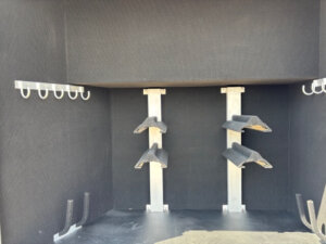 A black, carpeted storage compartment in the Bloomer 4H Trainer with metal brackets, angled shelves in the center, hooks on both walls, and padded holders attached to the floor. -Stillwater Trailer Sales