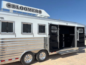 A silver Bloomer 4H Trainer horse trailer with multiple windows and an open side door revealing tack storage compartments, parked on a gravel surface under a clear blue sky. -Stillwater Trailer Sales