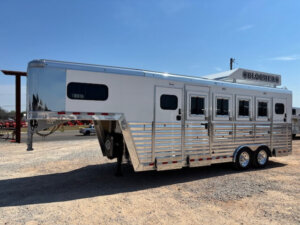 A large, silver Bloomer gooseneck horse trailer with multiple windows and vents is parked on a gravel lot under a clear blue sky. The trailer features dual axles and aluminum siding, perfect for a 4H Trainer or Trainer 549 needs. -Stillwater Trailer Sales