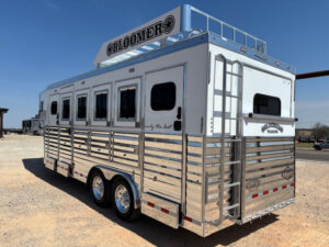 A large silver Bloomer 4H horse trailer with multiple windows, ventilation slats, and a ladder at the back, parked on gravel under a clear blue sky. The trailer features prominent BLOOMER branding on the top and side. -Stillwater Trailer Sales
