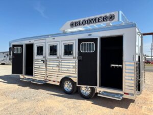 A silver Bloomer 4H Trainer horse trailer with three open doors, side windows with bars, and a rooftop rack, parked on gravel under a clear blue sky. -Stillwater Trailer Sales