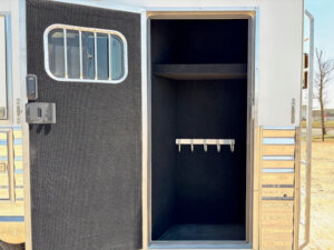 Open compartment of a Bloomer 4H Trainer horse trailer with black interior, a top shelf, and five hooks for hanging equipment. The door features a small barred window, and the silver trailer exterior has horizontal ridges. -Stillwater Trailer Sales