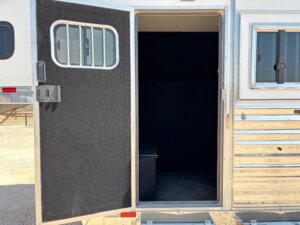 Open metal door on a Bloomer 4H trailer reveals a small, dark, carpeted interior space with a bench against the wall. The door has a barred window, and sunlight is visible outside. -Stillwater Trailer Sales