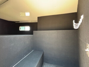 Interior of a small, black, padded Bloomer 4H Trainer room with a bench, partial divider wall, small window, and wall hooks; overhead lighting highlights the compact, minimalist design. -Stillwater Trailer Sales