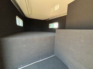 Interior view of an empty, carpeted Bloomer 4H Trainer horse trailer with a raised sleeping platform, black walls, and three rectangular windows with bars letting in daylight. -Stillwater Trailer Sales