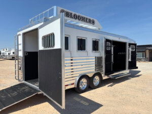 A white Bloomer 4H Trainer horse trailer with a ramp and open doors is parked on a dirt lot. Featuring multiple windows, metal trim, and two axles, the trailer sits under a clear sky with other trailers visible in the background. -Stillwater Trailer Sales