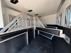 The image shows the interior of a Bloomer 4H horse trailer with divided stalls, padded dividers, and windows for ventilation and light. The black rubberized flooring ensures safety for your horses during transport. -Stillwater Trailer Sales