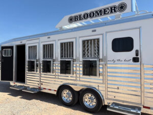 A shiny silver Bloomer 4H Trainer horse trailer with several windows and vents, parked on gravel under a clear blue sky. The logo BLOOMER and the phrase only the best are visible on the trailer. -Stillwater Trailer Sales