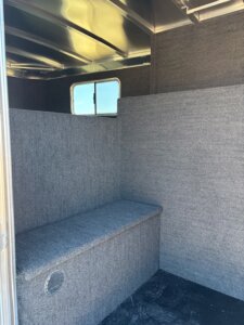 Interior of a trailer with gray carpeted walls and bench, black textured floor, and a small window showing blue sky&mdash;designed for the Elite 6 Horse line from Horse Smart Tack. -Stillwater Trailer Sales
