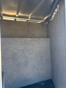 A small, empty room with gray carpeted walls, a metal ceiling, and a row of Smart Tack hooks on the upper right wall. The space appears enclosed and well-lit from above&mdash;ideal for organizing elite horse tack. -Stillwater Trailer Sales