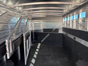 Interior view of an empty Elite 6 aluminum livestock trailer with black rubber flooring, slatted vents for ventilation, and a partition gate. Sunlight casts shadows through the side vents onto the floor&mdash;Horse Smart Tack quality. -Stillwater Trailer Sales
