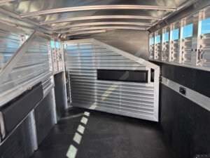 Interior of an empty Elite 6 Horse aluminum livestock trailer with ribbed metal walls, black padded bumpers, open air slots on the right, and a partially open partition gate in the center. Tack #23223 and Horse Smart Tack features included. -Stillwater Trailer Sales