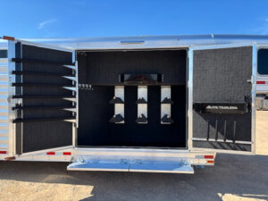 Open trailer door revealing an organized Horse Smart Tack room with multiple saddle racks, bridle hooks, and storage compartments against black interior walls, set outdoors under a blue sky. Perfect for storing your Elite 6 gear. -Stillwater Trailer Sales