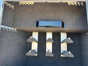 A carpeted storage compartment in the Elite 6 Horse trailer with multiple metal hooks on the walls, a black rectangular box on the back wall, and three metal supports with carpeted pads on the floor&mdash;part of Horse Smart Tack #23223. -Stillwater Trailer Sales