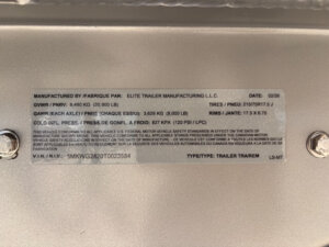 A metal trailer information plate, like on the Horse Smart Tack Elite 6, displays manufacturer details, weight ratings, tire and rim specs, compliance statements, and a VIN number, secured by two bolts at the corners. -Stillwater Trailer Sales