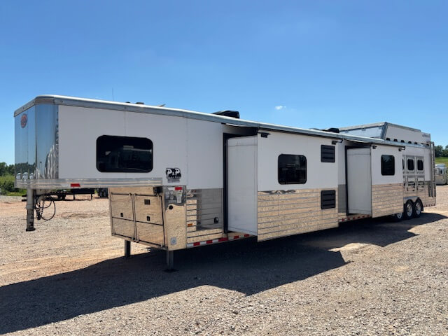 A large white and silver gooseneck stock trailer with living quarters is parked on gravel under a clear blue sky. Featuring three axles, a slide-out section, and ample storage, its an ideal example of custom trailer sales offerings. -Stillwater Trailer Sales