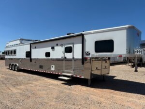 A large white and silver gooseneck horse trailer with multiple windows and three axles, perfect for Trailer sales or Living Quarters custom options, is parked on a gravel lot under a clear blue sky. -Stillwater Trailer Sales