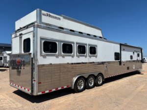 A large silver Custom Show Trailer with multiple windows and three axles is parked on gravel under a clear blue sky. The trailer features a polished lower exterior and prominent branding on the upper front section. -Stillwater Trailer Sales