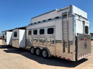 A large silver horse trailer with multiple windows and three axles is parked on gravel. Ideal for those seeking new and used trailers, it features slide-out sections and a raised roof area under a clear blue sky. -Stillwater Trailer Sales