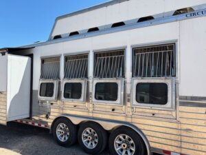 A silver livestock or horse trailer with three axles is parked on gravel. This Custom Show Trailer features five windows with metal bars, ventilation above, and an open side door. The sky is clear and blue. -Stillwater Trailer Sales