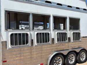 A close-up view of the side of a silver horse trailer highlights four windows with metal bars above four small doors. Perfect for horse and livestock trailer sales, this two-axle trailer is parked on a sunny day. -Stillwater Trailer Sales