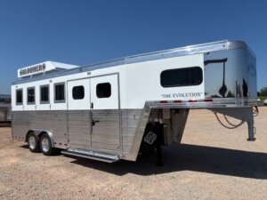 A silver and white gooseneck horse trailer with three windows and two doors is parked on gravel under a clear blue sky. Perfect for those seeking quality horse and livestock trailer sales, it features BLOOMER and THE EVOLUTION written on it. -Stillwater Trailer Sales