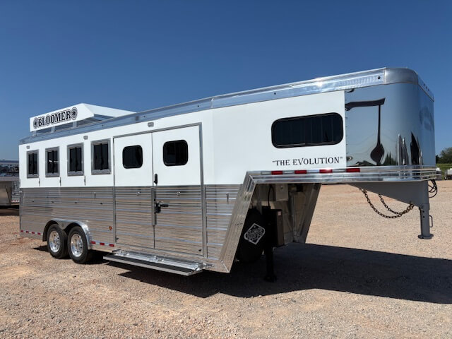 A silver and white gooseneck horse trailer labeled THE EVOLUTION and BLOOMER is parked on a gravel lot under a clear blue sky. This top choice in stock trailers features multiple windows, dual axles, and is available in our trailer sales inventory. -Stillwater Trailer Sales
