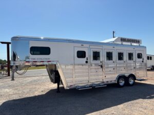 A silver, multi-compartment horse trailer with “The Evolution” and “Bloomer” branding is parked on a gravel lot under a clear blue sky. This Custom Show Trailer features windows and separate doors for each compartment. -Stillwater Trailer Sales