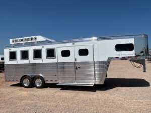 A silver and white gooseneck horse trailer with four windows and two axles is parked on a gravel lot under a clear blue sky. The Bloomer The Evolution is perfect for those seeking custom show trailers or quality new and used trailers. -Stillwater Trailer Sales