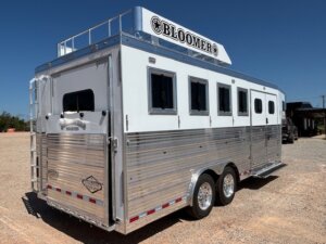 A white and silver Bloomer-brand horse trailer with four windows, two axles, and a roof rack, parked on a gravel surface under a clear blue sky—perfect for those seeking new and used trailers or exploring top Stock Trailers options. -Stillwater Trailer Sales