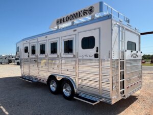 A silver Bloomer horse trailer with four windows, dual axles, a roof ladder, and a rooftop sign is parked on gravel under a clear blue sky—perfect for those seeking quality horse and livestock trailer sales. -Stillwater Trailer Sales