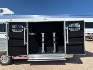 A Custom Show Trailer with both rear doors open reveals an interior tack room featuring saddle racks and bridle hooks. The trailer is parked on gravel under a clear blue sky—explore new and used trailers with Living Quarters custom options. -Stillwater Trailer Sales