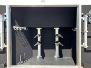 Open tack room compartment of a horse and livestock trailer featuring two vertical saddle racks, bridle hooks on both side walls, and a boot jack on the floor. Explore new and used trailers with interiors lined in dark carpeting. -Stillwater Trailer Sales
