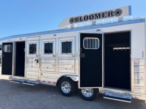 A silver Bloomer Stock Trailer with multiple open doors and windows, parked on gravel under a clear blue sky. The shiny metal siding and two visible wheels highlight its quality—perfect for those seeking new and used trailers at top trailer sales. -Stillwater Trailer Sales