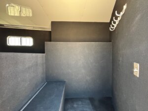 Interior of a small, black-walled compartment in a Living Quarters custom trailer with a window, metal hooks, seating ledge, and outlet. The space appears clean and minimalistic—ideal for modern horse and livestock trailer sales. -Stillwater Trailer Sales