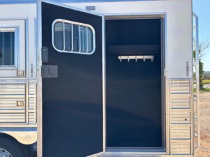 A horse trailer with its side door open, revealing an empty interior with a row of hooks mounted on the wall. Perfect for those seeking Custom Show Trailers or Stock Trailers, the door features a small window with metal bars. -Stillwater Trailer Sales